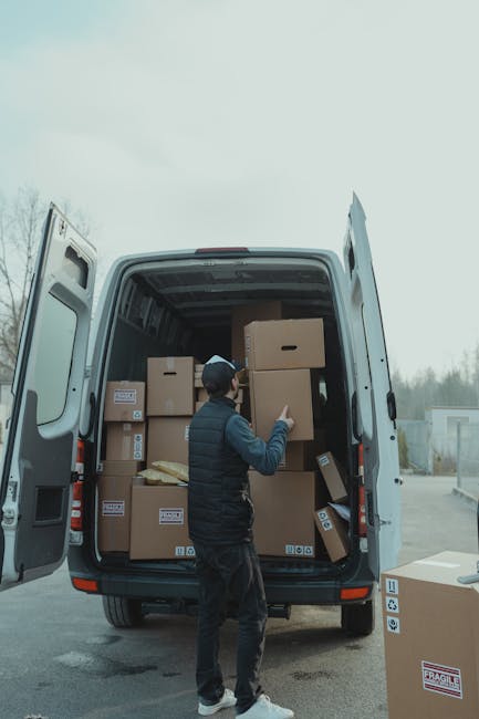 A grey moving van parked on an unpaved surface with its side door open, revealing a wooden wardrobe and various boxes inside. The van is positioned in an outdoor area overlooking a cityscape during du