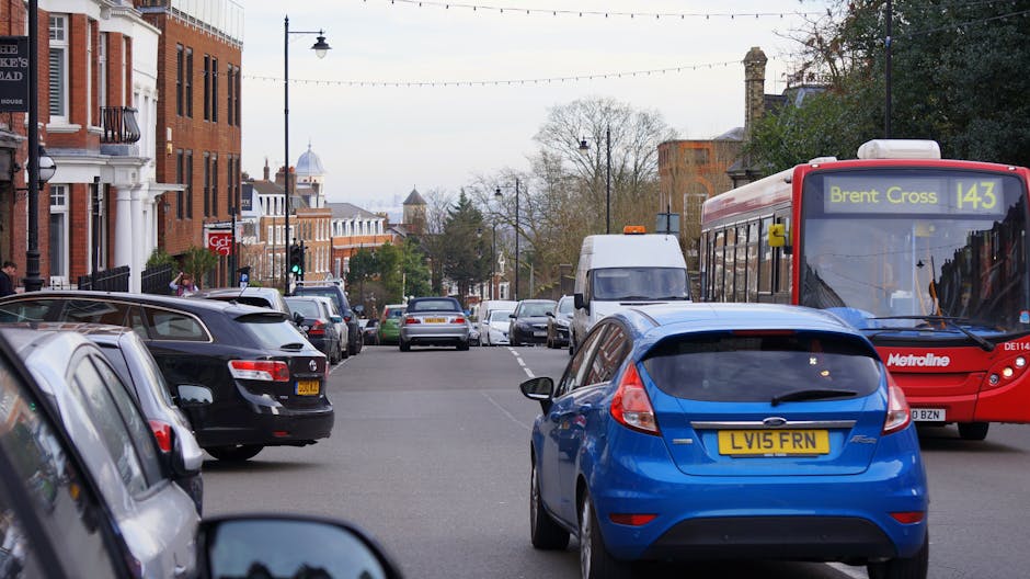 A city street scene with multiple parked cars along both sides, including a black, silver, and white vehicle, with a blue Ford Focus with the registration LVI5 FRN in the foreground. A red Metroline bus displaying route 143 towards Brent Cross is visible on the right side, with a white delivery van further down the street. The background features a row of brick and white residential buildings, some with balconies and decorative facades, under an overcast sky, with street lamps and leafless trees indicating a late autumn or winter season. The scene captures a typical urban environment suitable for house removals and furniture transport, with the placement of vehicles and urban infrastructure illustrating the logistical considerations involved in home relocation and packing and moving processes, as managed by services like Man with Van Crook Log.