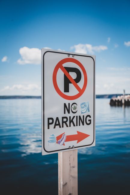A vertical rectangular traffic sign mounted on a wooden post situated on a paved area near a body of water under a clear sky with some clouds. The sign features a large black 'P' with a red circle and diagonal line crossing it, indicating no parking, along with the words 'NO PARKING'. The sign also has a small green cannabis leaf icon, a blue water droplet, and a red and white striped object, possibly a boat or life jacket, at the bottom. An orange arrow points to the right, indicating the direction for parking restrictions. In the background, there is calm water with a distant shoreline, a breakwater made of large rocks extending into the water, and partly cloudy sky. The scene suggests a coastal or lakeside location, possibly near residential or leisure properties where moving and transport logistics, such as house removals, might be relevant. Man with Van Crook Log's professional removal services could assist with planning home relocations or furniture transport in the area.
