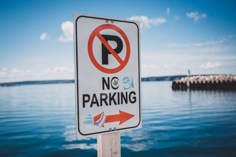 A vertical rectangular sign with a white background and a black border displaying a red circle with a slash over a black 'P', indicating no parking. Below, the words 'NO PARKING' are printed in bold black letters. There are two stickers on the sign: one with a green cannabis leaf and another with a stylized face mask. An orange arrow points to the right at the bottom of the sign, which also features small images of a cupcake and a high-heeled shoe. The sign is mounted on a wooden post situated outdoors near a body of water, with a blue sky, a few clouds, and a distant breakwater visible in the background. The context suggests the sign is likely related to parking restrictions in a coastal or waterfront area, and it is positioned in the vicinity of house removals or moving services such as those provided by Man with Van Crook Log, possibly to indicate parking rules during moving or loading activities.