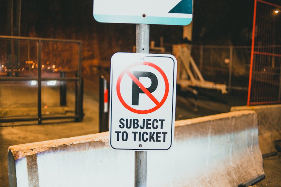 A vertical rectangular traffic sign mounted on a wooden post situated on a paved area near a body of water under a clear sky with some clouds. The sign features a large black 'P' with a red circle and diagonal line crossing it, indicating no parking, along with the words 'NO PARKING'. The sign also has a small green cannabis leaf icon, a blue water droplet, and a red and white striped object, possibly a boat or life jacket, at the bottom. An orange arrow points to the right, indicating the direction for parking restrictions. In the background, there is calm water with a distant shoreline, a breakwater made of large rocks extending into the water, and partly cloudy sky. The scene suggests a coastal or lakeside location, possibly near residential or leisure properties where moving and transport logistics, such as house removals, might be relevant. Man with Van Crook Log's professional removal services could assist with planning home relocations or furniture transport in the area.