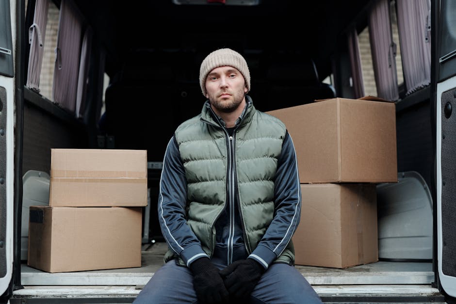 Two men are engaged in a home relocation process outside a residential property. One man, standing beside an open moving van, is wearing a baseball cap, dark jacket, and white sneakers, and appears to