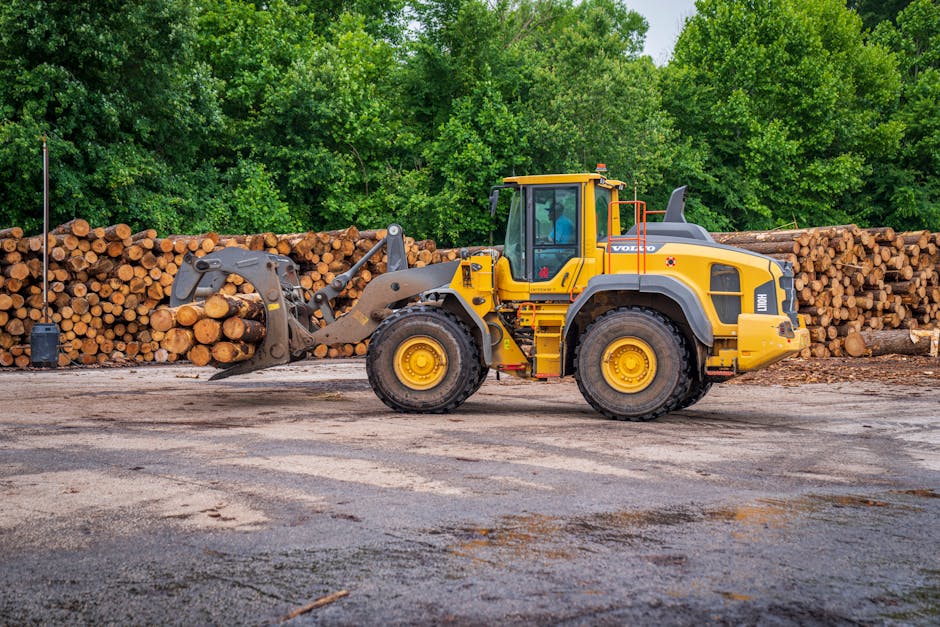 A yellow Volvo front loader with a cab containing a seated operator is positioned on an asphalt surface, engaged in stacking or moving large logs of wood. The logs, which are uniform in diameter, are neatly arranged in two horizontal piles behind the machinery, extending across the background. The logs are in natural wood tones, with some showing bark, and are stacked on a dirt and gravel area adjacent to the asphalt. The front loader’s hydraulic arm and grapple are gripping a small section of logs, indicating active loading or handling in a wood storage yard. Surrounding the yard is dense greenery with leafy trees and shrubs, suggesting an outdoor, rural, or semi-industrial environment. The scene captures a typical setting involved in the transportation or preparation of wooden materials, which may be part of a house removal or bulk material transport process. Man with Van Crook Log occasionally operates vehicles like this during home relocation and furniture transport, supporting efficient packing and moving logistics.