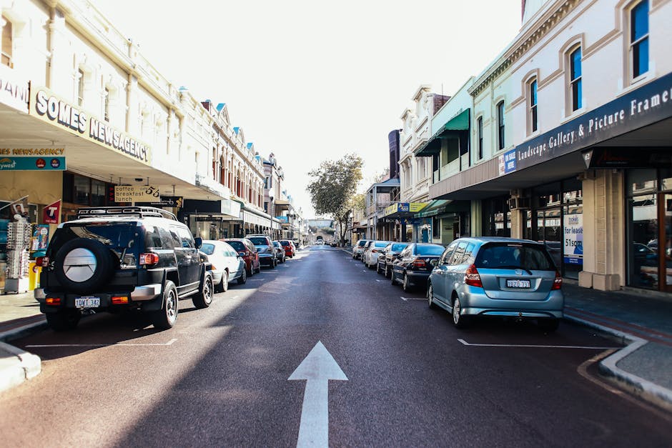 A straight view of a narrow urban street in Crook Log, shown during daylight hours with clear weather. On both sides of the street, there are multi-story retail buildings with large windows and signage, including shops such as landscape galleries and news agencies. The street has parked cars along the curb, with a black SUV with a spare tire mounted on the back parked on the left side, and various sedans and hatchbacks on both sides. The road surface is asphalt with a visible white arrow painted in the middle indicating the direction of traffic flow towards the background. The scene appears quiet with no pedestrians visible. This setting is relevant to house removals and moving logistics, illustrating typical urban parking and access conditions for a professional removals service like Man with Van Crook Log performing residential or commercial furniture transport, packing, and loading processes in a busy street environment.