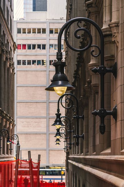 A series of ornate black metal street lamps with decorative scrollwork are mounted along the side of a historic building in an urban setting, with one lamp featuring a glowing yellow bulb. In the background, there is a modern multi-storey office building with numerous windows, contrasting with the older architectural style of the foreground. The street appears narrow, and the lamps extend outward from the building façade. Part of a red safety barrier or construction fencing is visible along the pavement, indicating ongoing work or access restrictions. The scene is outdoors during daylight, with natural light illuminating the intricate details of the street lighting and surrounding architecture. This image captures elements typical of a city street involved in a home relocation or moving process, where careful access and parking considerations are important. As part of the services provided by Man with Van Crook Log, such street fittings are relevant to the logistical planning of furniture transport and packing during house removals in the Crook Log area.