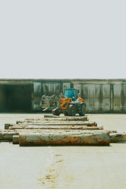 A heavy-duty flatbed truck loaded with large logs of varying lengths and diameters parked on a street with a clear blue sky overhead. The logs, some with visible bark and others partially stripped, ar