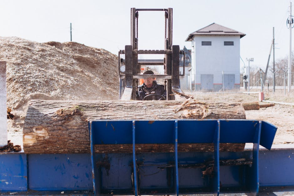 A close-up view of a worker operating a forklift at an outdoor site, lifting a large wooden log into a blue industrial skip. The forklift is positioned behind the log, with the operator seated inside, partially visible, focused on the task. The background features a pale building, possibly a warehouse or storage facility, with a pitched roof, small windows, and ventilation openings, along with some fencing, utility poles, and trees indicating an industrial or construction environment. The ground is covered with gravel and dirt, typical of a site where furniture and materials might be loaded or unloaded during house removals or home relocation activities. The scene illustrates heavy-duty equipment used in furniture transport and packing during a move, highlighting the logistics of loading large items for transport with professional assistance, such as Man with Van Crook Log.